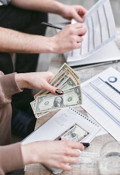 Two individuals counting dollar bills and reviewing financial documents indoors.