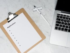 A clean and organized office desk setup featuring a laptop, clipboard with application form, and pen.
